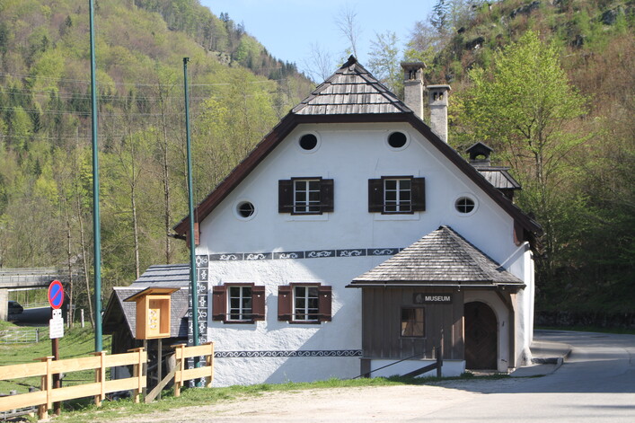 Ältester „Paarhofverband“ im Salzkammergut mit Wohnhaus, Stallstadel sowie angeschlossener Sägerei, Mühle, Gipsstampfe und Backstube. Ein kleines Museum in einem traditionellen weißen Haus mit Holzschindeldach. Fensterläden in dunklem Braun, dekorative Muster an den Wänden. Im Hintergrund grüne Hügel und Bäume, ein Weg führt am Gebäude vorbei.