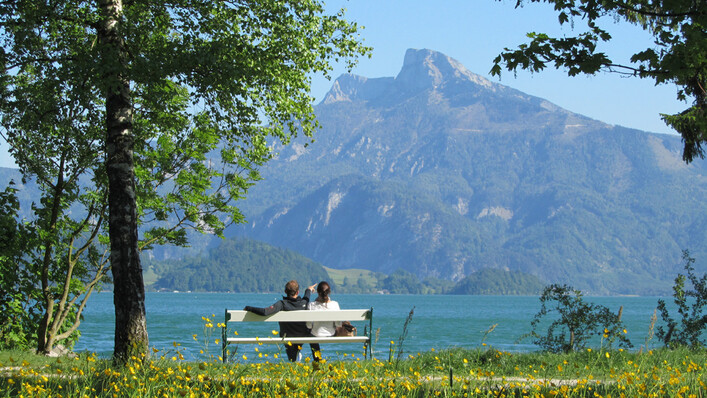 Personen sitzen auf einer Bank, vor ihnen der See und die Berge