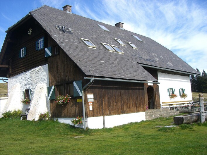 Ein traditionelles Holzhaus mit einem steilen Dach und bunten Fenstern. Das Gebäude steht in einer grünen Wiesenlandschaft unter einem blauen Himmel.