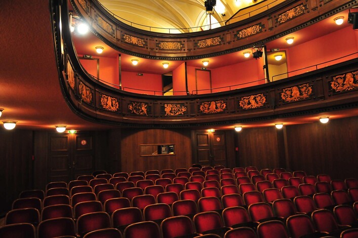 Ein gemütliches Theater mit roten Sitzen und einem eleganten Balkon. Die warmen Lichter schaffen eine einladende Atmosphäre.