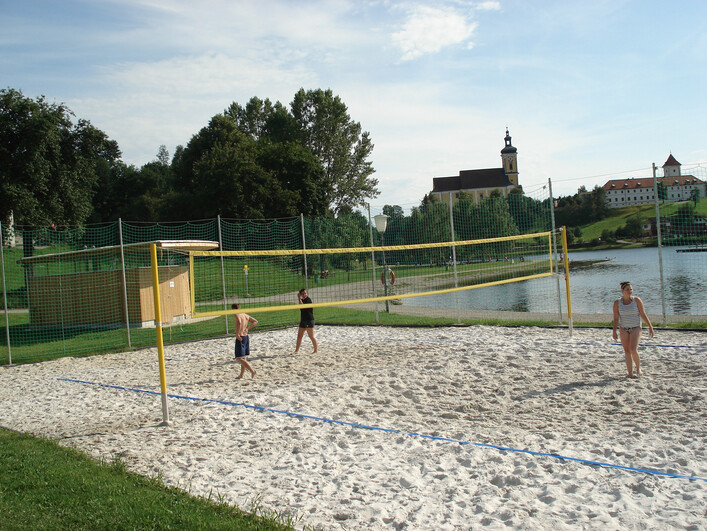 Beach-Volleyballplatz-Badesee in Waldhausen im Strudengau: Eine Strandvolleyballanlage an einem See oder Fluss. Im Vordergrund sieht man einen Sandplatz mit Volleyballnetz, dahinter erstreckt sich eine Parklandschaft mit Bäumen und in der Ferne ist ein Kirchturm zu erkennen.
