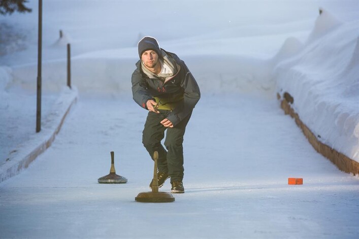 Ein Mann spielt Curling auf einer eisigen Bahn. Die Umgebung ist schneebedeckt und es ist helles, kaltes Wetter.