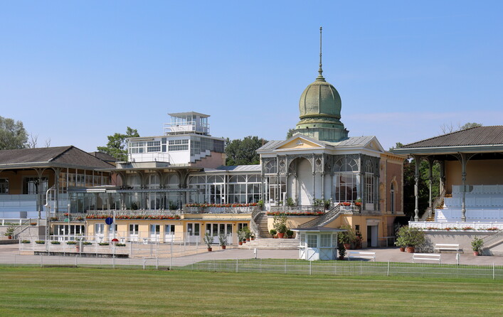 Die Hof-Tribüne der Galopprennbahn Freudenau im 2. Wiener Gemeindebezirk Leopoldstadt.Die Galopprennbahn ist eine in der Freudenau 1839 eröffnete Anlage. 1858 findet in Gegenwart von Kaiser Franz Joseph die Einweihung von 2 Tribünen statt, die nach Entwürfen des Ringstraßen-Architekten Carl Hasenauer von seinem Bruder, dem Hofzimmermeister Christoph Hasenauer, errichtet wurden. 1870 erfolgte die Einweihung der „Hof-Tribüne“, die aus der „Hof-Loge“, der Clubloge und der „westlichen“ Tribüne besteht, und vom Budapester Architekten Adolf Feszty (1846–1900) entworfen wurde. 1883 zerstörte ein Brand teilweise die Tribünen. Für die Wiederinstandsetzung wurde der Architekt Josef Drexler beauftragt. Im Anschluss wurden nach seinen Plänen bis 1887 auch noch das Totalisator-Gebäude, das Verwaltungs- sowie Stallgebäude errichtet. 1967 kaufte der Staat um 7,6 Mio. Schilling die Gebäude auf dem Rennplatz. Von 1983 bis 1986 fand eine aufwendige Renovierung der Tribünenanlage statt. 1991 Gründung der IRM-Interrace Rennbahn Management Ges.m.b.H., die die Anlage mit den Stallungen „verwaltet“. De facto finden keine Rennen mehr statt. Die Hoftribüne wird für Events aller Art genutzt und die Rasenfläche innerhalb der 2.800 km langen Rennbahn als Golfplatz.