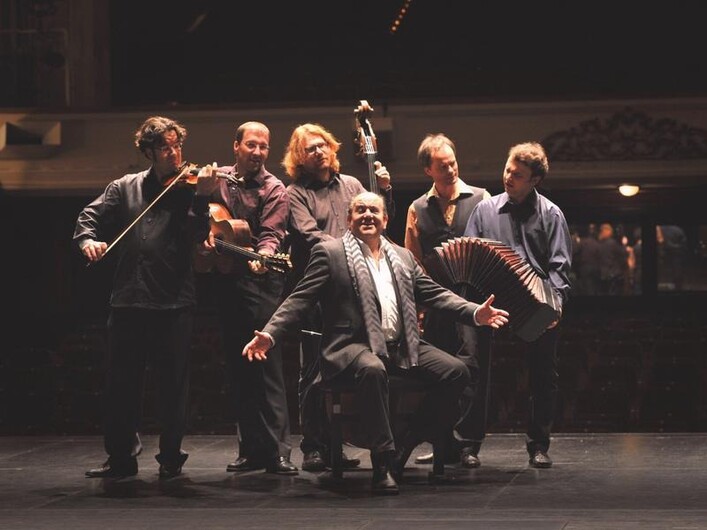 Eine Gruppe von sechs Musikern auf einer Bühne. Sie spielen verschiedene Instrumente und haben Spaß beim Musizieren.