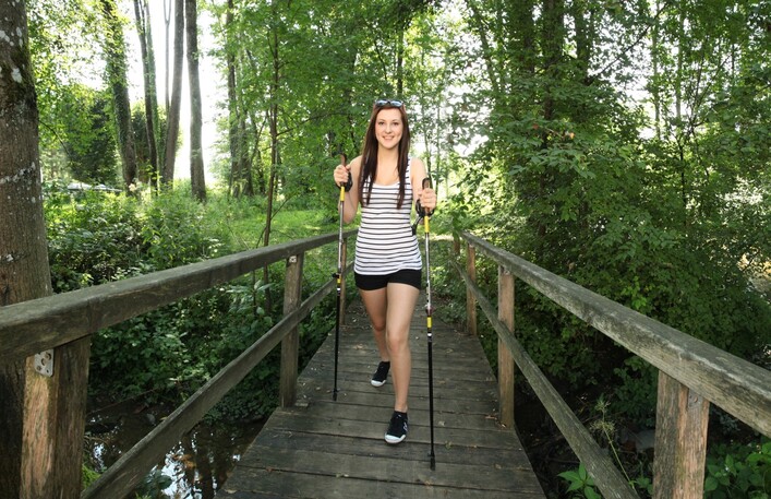 Bewegungsarena Mattigtal in Mattighofen: Eine junge Frau in sportlicher Kleidung steht auf einer Holzbrücke in einem dichten Wald. Sie hält Wanderstöcke in den Händen und lächelt in die Kamera. Der Wald um sie herum ist grün und üppig.