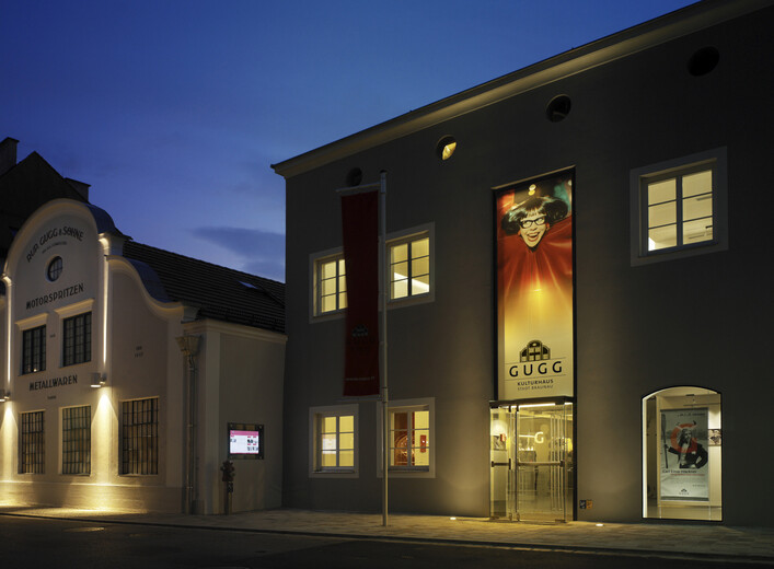 Gugg Kulturhaus in Braunau am Inn: Ein mehrstöckiges Gebäude mit beleuchteten Fenstern und einer Eingangstür. An der Fassade befindet sich ein großes Schild mit dem Namen "Gugg". Es scheint sich um ein kulturelles Zentrum oder eine ähnliche Einrichtung zu handeln.