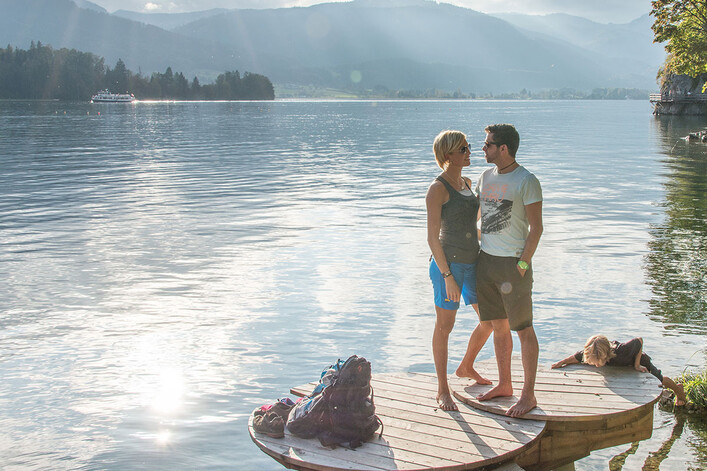 Wolfgangsee in St. Wolfgang im Salzkammergut: Das Bild zeigt ein junges Paar, das auf einem Holzsteg an einem See stehend, die malerische Landschaft mit Bergen und einem Ausflugsschiff im Hintergrund betrachtet. Die Umgebung vermittelt eine friedliche und entspannende Atmosphäre.