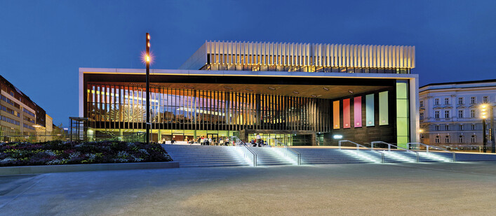 Musiktheater am Volksgarten, Linz: Modernes Theatergebäude mit beleuchteter Fassade und vertikalen Lamellen in der Abenddämmerung.