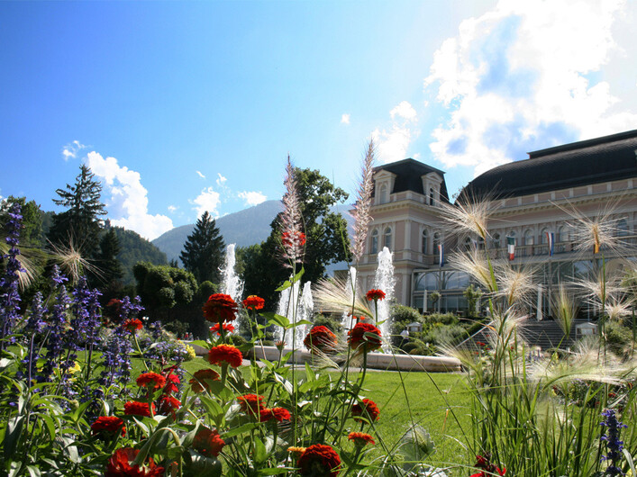 Kurpark Bad Ischl in Bad Ischl: Das Bild zeigt ein prächtiges Gebäude mit traditioneller Architektur, umgeben von einem wunderschönen Garten voller Blumen und Pflanzen. Im Vordergrund befinden sich bunte Blumen, während im Hintergrund Berge zu sehen sind.
