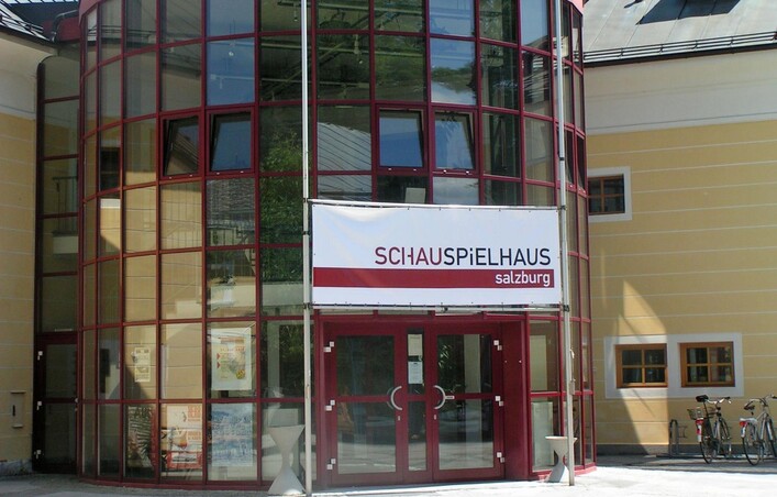 <p>Ein modernes Gebäude mit rot umrandetem Glas und einer großen Eingangstür. Über dem Eingang hängt ein Schild mit dem Schriftzug "SCHAUSPIELHAUS Salzburg".</p>