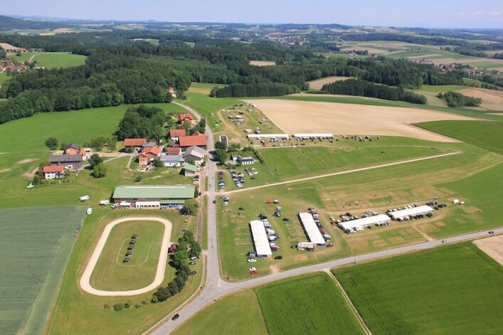 Islandpferdehof Lichtegg in Andorf: Eine ländliche Umgebung mit mehreren Gebäuden und Parkplätzen, umgeben von Feldern und Waldgebieten. Es gibt eine kreisförmige Struktur, die möglicherweise eine Rennstrecke oder Sportstätte darstellt. Insgesamt eine idyllische Landschaft mit einer Mischung aus landwirtschaftlichen Flächen und bewaldeten Gebieten.