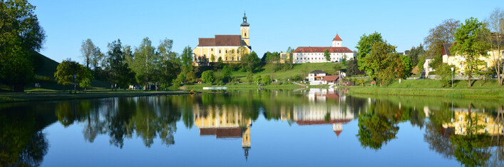 Badesee Waldhausen im Strudengau in Waldhausen im Strudengau: Eine mittelalterliche Burg mit einem hohen Turm und roten Dächern, die sich an einem See mit klarem Wasser und Bäumen am Ufer widerspiegelt. Die Landschaft ist von grünen Wäldern und Hügeln umgeben, was eine malerische und friedliche Szene ergibt.