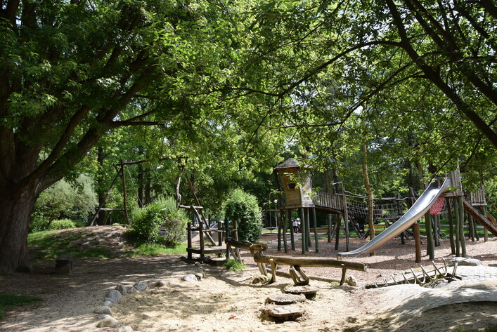 Freizeitgelände Vöcklabruck in Vöcklabruck: Ein grüner, waldähnlicher Erholungsbereich mit einem Spielplatz. Große, schattenspendende Bäume rahmen einen Pfad ein, der zu Spielgeräten wie einer Rutschbahn und Kletterstrukturenführt. Der Boden ist mit Kies bedeckt, was einen natürlichen Charakter verleiht. Es wirkt wie ein einladender, waldiger Ort zum Spielen und Entspannen.