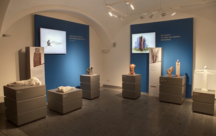 Daringer Kunstmuseum Aspach in Aspach: Ein Ausstellungsraum mit mehreren Kunstwerken und Objekten. Es sind verschiedene Skulpturen und Reliefs zu sehen, die menschliche Figuren und Formen darstellen. Die Werke sind auf Podesten und Regalen platziert und vor einem blauen Wandhintergrund präsentiert. Die Beleuchtung ist hell und fokussiert die Ausstellungsstücke. Insgesamt vermittelt der Raum einen ruhigen und musealen Eindruck.