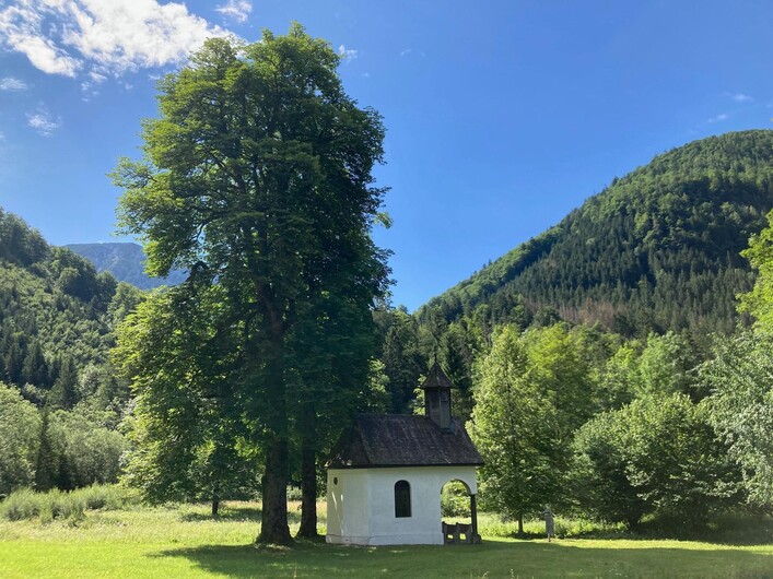 Eine kleine Kapelle umgeben von hohen Bäumen und grünen Wiesen. Im Hintergrund erheben sich majestätische Berge unter einem blauen Himmel.