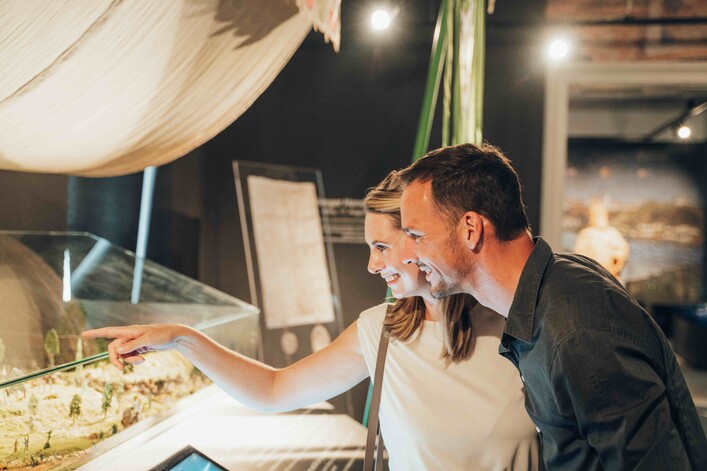 Museum 1212 Enns in Enns: Ein junges, freundliches Paar steht an einer Kasse und scheint ein Produkt auszuwählen. Die Frau deutet mit ihrer Hand auf etwas auf der Anzeige, während der Mann neben ihr lächelt. Hinter ihnen ist eine Sitzecke mit gemütlicher Beleuchtung und dekorativen Elementen zu sehen.