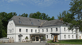 Sturmmühle in Saxen: Ein charmantes Gebäude mit traditioneller Architektur, umgeben von Bäumen und Grünflächen. Die Fassade ist hell gestrichen, mit mehreren Stockwerken und Dachgauben. Am Eingang ist eine Terrasse mit Sitzmöbeln und dekorativen Elementen.