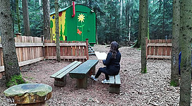 Waldgeschichten in Roßbach: Ein grünes Holzhäuschen inmitten eines Waldes, davor ein Holzbank, auf der eine Person sitzt. Um das Häuschen herum sind weitere Holzelemente, die vermutlich zum Spielen oder Verweilen gedacht sind.