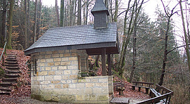 Heil-Bründl-Kapelle in St. Radegund: Eine rustikale Holzhütte mit einem spitzen Dach steht inmitten eines bewaldeten Gebiets. Stufen führen zu einer überdachten Veranda, die zum Betreten der Hütte einlädt. Die Umgebung ist von Bäumen und Sträuchern umgeben, was eine idyllische und gemütliche Atmosphäre schafft.