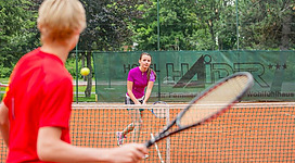 Zwei Personen spielen Tennis auf einem Sandplatz, im Vordergrund ein Spieler in rotem Shirt, im Hintergrund eine Spielerin in lila Shirt am Netz.