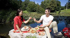 Ein Paar genießt ein Picknick am Ufer eines ruhigen Teiches. Sie teilen sich frisches Obst und lachen in der schönen Natur.
