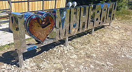 Ein glänzendes Schild mit der Aufschrift "I ❤️ Mühlbach" steht in der Natur. Im Hintergrund sind Bäume und eine malerische Landschaft zu sehen.