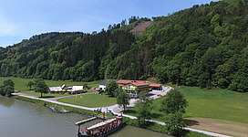 Gasthof in der Exlau in Kirchberg ob der Donau: Eine idyllische Landschaft mit einem Hotelkomplex am Ufer eines Flusses oder Sees. Der Hotelkomplex besteht aus mehreren Gebäuden in traditioneller Architektur, umgeben von üppiger Vegetation und Hügeln im Hintergrund. Auf dem Wasser befindet sich ein historisch anmutender Bootssteg oder Steg.