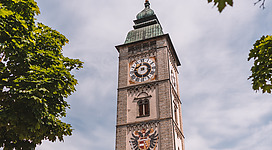 Stadtturm in Enns: Ein hoher Turm mit einer kunstvoll verzierten Fassade thront inmitten eines malerischen Stadtbilds. Die Uhr am Turm ist deutlich erkennbar, darüber befindet sich ein ornamentierter Aufbau. Grüne Baumkronen rahmen den Turm und verleihen dem Gesamtbild eine harmonische Atmosphäre.