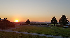 Römerbad Villa Weirading in Altheim: Eine idyllische Landschaft mit grünen Wiesen, Bäumen und einem malerischen Sonnenuntergang am Horizont. Das Gelände wirkt friedlich und ruhig, mit sanften Hügeln und einem Weg, der durch die Szene führt.