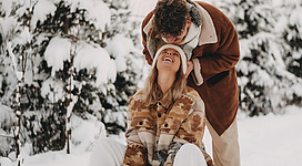 Ein Paar sitzt im Schnee und lacht miteinander. Die Umgebung ist winterlich mit schneebedeckten Bäumen.