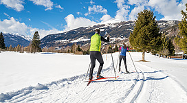 Zwei Personen gehen im Schnee Ski-Langlauf. Im Hintergrund sind schneebedeckte Berge und ein blauer Himmel mit wenigen Wolken zu sehen.
