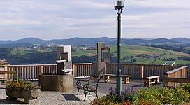 Sauwald-Panoramastraße in Vichtenstein: Eine überdachte Terrasse mit Blick auf eine bergige Landschaft. Eine Straßenlaterne und Bänke laden zum Verweilen ein. Zwischen Pflanzen und Blumen befindet sich ein Brunnen. Insgesamt bietet die Szene einen idyllischen und gemütlichen Eindruck.