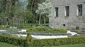 Burggarten Obernberg in Obernberg am Inn: Eine große Burg oder ein historisches Gebäude mit verwitterter Fassade, umgeben von blühenden Bäumen und einer gepflegten Gartenanlage. Im Vordergrund befinden sich Beete mit verschiedenen Blumen und Sträuchern. Vor dem Gebäude ist ein geschwungener Weg angelegt, der von einem gusseisernen Tor begrenzt wird.