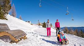 Eine Familie genießt einen sonnigen Tag im Schnee. Im Hintergrund sind Skilifte und verschneite Berge zu sehen.