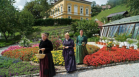 Drei Frauen in traditioneller Tracht gehen durch einen blühenden Garten. Im Hintergrund ist ein hübsches, gelbes Haus und eine Gewächshaus zu sehen.