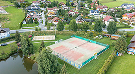 Eine Luftaufnahme eines Tennisplatzes umgeben von schönen Wohnhäusern und grünen Wiesen. Im Hintergrund sieht man einen kleinen See und bewaldete Hügel.