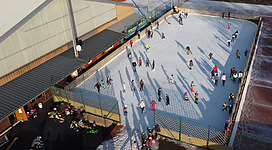 Eislaufplatz Schwanenstadt in Schwanenstadt: Eine große Eisfläche mit zahlreichen Menschen, die darauf Schlittschuh laufen oder andere Aktivitäten ausüben. Um die Eisfläche herum befinden sich Gebäude und Einrichtungen, die vermutlich Teil einer Wintersportanlage sind.