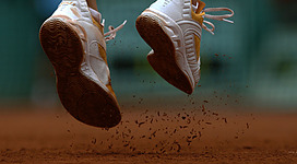 Tennisplätze in Grein: Das Bild zeigt die vom Boden hochfliegenden Füße einer Person, die auf einer sandigen Oberfläche läuft. Die Schuhe haben eine markante Sohle mit deutlich sichtbarem Profil. Staub und kleine Partikel spritzen in der Luft umher, was den dynamischen Eindruck der Bewegung verstärkt.