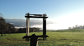 Burgblick Schauern in St. Aegidi: Eine Holzstruktur auf einer grünen Wiese, umgeben von bewaldeten Hügeln und grauem Morgennebel. Die Struktur besteht aus einem Querbalken, der von zwei vertikalen Stangen getragen wird. Die Szene vermittelt einen friedlichen, ländlichen Charakter.