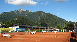 Tennisclub St. Gilgen in St. Gilgen: Das Bild zeigt einen Tennisplatz mit mehreren Spielern in einer bergigen Umgebung. Im Hintergrund sind große Felsen und dicht bewaldete Berghänge zu sehen, über denen sich ein blauer Himmel mit weißen Wolken erstreckt.