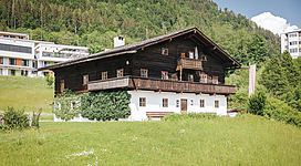 Ein traditionelles Holzhaus mit einem Balkon steht in einer grünen Wiese. Im Hintergrund sind moderne Gebäude und eine bewaldete Hügelkette zu sehen.