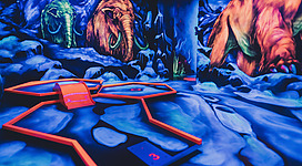 Leuchtend buntes Indoor-Minigolf mit fluoreszierenden Farben und leuchtenden Mammut-Wandbildern in einer fantasievollen Höhlenlandschaft.