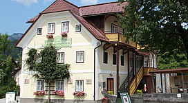 Auf diesem Foto siehst du das Heimat- und Landlermuseum in Bad Goisern, nach der Generalsanierung 2014. Es ist ein typisches Salzkammergut Haus, die Fassade ist gelb gestrichen und das Dach ist dunkelrot. An der Vorderseite ist ein grünes Schild mit der Aufschrift des Museums angebracht. Zusätzlich befindet sich direkt an der Fassade ein Spalierbaum, welcher in unserer Gegend eine Besonderheit ist. Rechts ein Stiegenaufgang und die typische Veranda in gelb und dunkelrot.