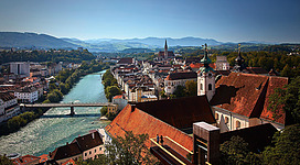 Eine malerische Stadtansicht mit einem fließenden Fluss und historischen Gebäuden. Im Hintergrund erstrecken sich die sanften Hügel und Berge unter einem blauen Himmel.