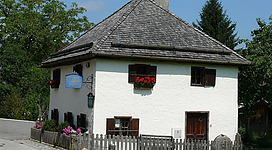 Ein traditionelles, zweigeschossiges Haus mit einem steilen Dach. Es hat Fenster mit Blumen und einen eingezäunten Vorgarten.
