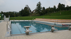 Freibad Ampflwang in Ampflwang im Hausruckwald: Ein Schwimmbad im Freien mit mehreren Becken. Auf einer Seite befinden sich erhöhte Holztribünen. Grüne Wiesen und Bäume umgeben das Schwimmbad. In der Ferne sind Gebäude zu sehen, die in die Landschaft eingebettet sind.