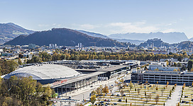 Ein moderner Veranstaltungsort mit einer großen Rasenfläche und Bäumen. Im Hintergrund sind Berge und eine Stadtlandschaft zu sehen.