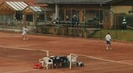 Zwei Personen spielen Tennis auf einem roten Platz. Im Hintergrund sind Gebäude und Zuschauer zu sehen.