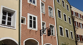 Heimathaus - Glockengießerhaus in Braunau am Inn: Ein mehrgeschossiges Gebäude mit Bögen und Fenstern an der Fassade. Es hat einen traditionellen architektonischen Stil mit Ziegeldach und Erkern. Vor dem Gebäude befinden sich Verkehrsschilder und Blumen auf dem Gehsteig, was einen malerischen Gesamteindruck vermittelt.