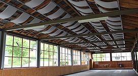 Asphaltbahnen Aubach in St. Martin im Mühlkreis: Ein großer Innenraum mit hoher Holzdecke, die an ein Wellendach erinnert. Große Fenster an den Wänden lassen viel Tageslicht herein. Der Boden ist mit Dielen belegt, und es sind einige Sitzmöbel zu sehen.